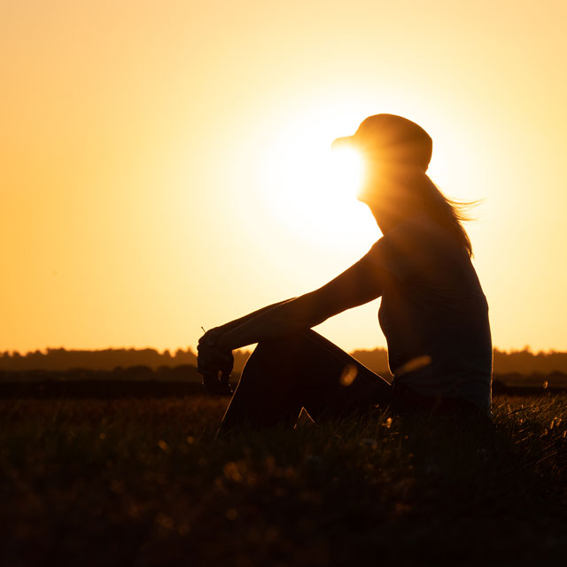 A silhouette of a woman wearing a ballcap, sitting in a field at sunset.